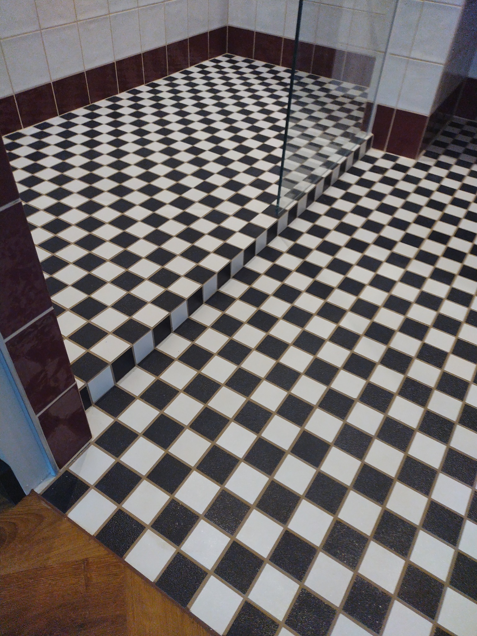 Ukończona podłoga łazienki wyłożona czarno-białą szachownicą, z widocznym fragmentem szklanej kabiny prysznicowej i brązowym pasem płytek przy ścianie.