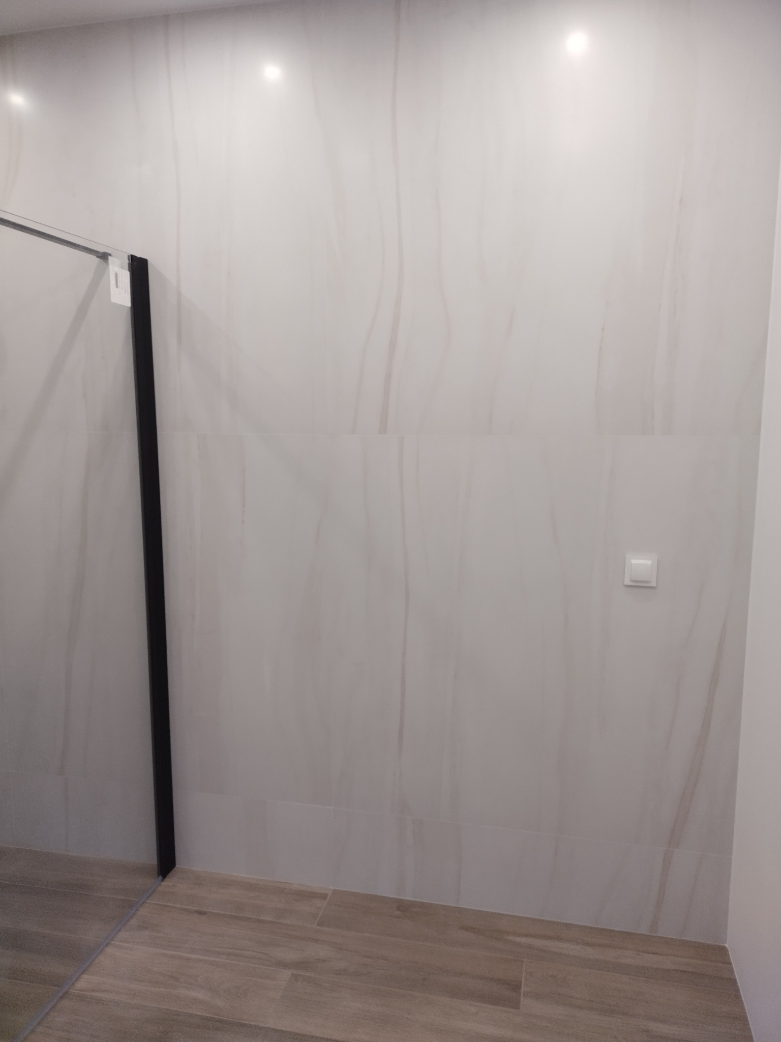 Wykończona łazienka z dużymi, jasnymi płytkami imitującymi marmur na ścianach, drewnopodobną podłogą i czarną ramą szklanej kabiny prysznicowej.