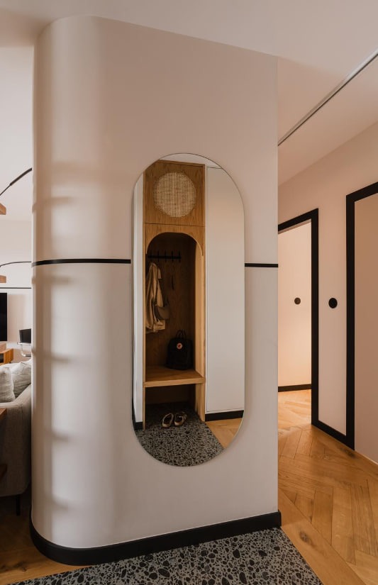 Nowoczesny przedpokój z owalnym lustrem w zabudowie, drewnianym panelem z rattanową plecionką, wieszakami na ubrania, półką, plecakiem i butami, wykończony listwami w kolorze czarnym.