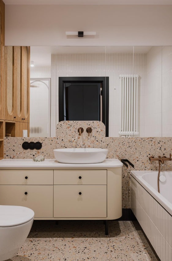 Nowoczesna łazienka z umywalką nablatową, beżową szafką z czarnymi gałkami, ścianą i podłogą wykończoną terazzo, czarną stolarką drzwiową i złotą armaturą