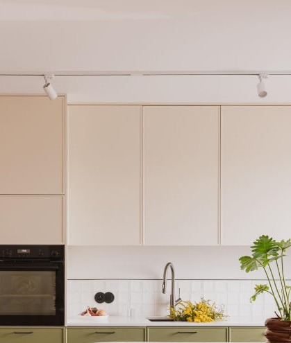 Jasna kuchnia z kremowymi szafkami górnymi i oliwkowymi dolnymi, oświetlenie szynowe, białe płytki nad blatem, nowoczesna bateria, roślina doniczkowa i owoce w misce.