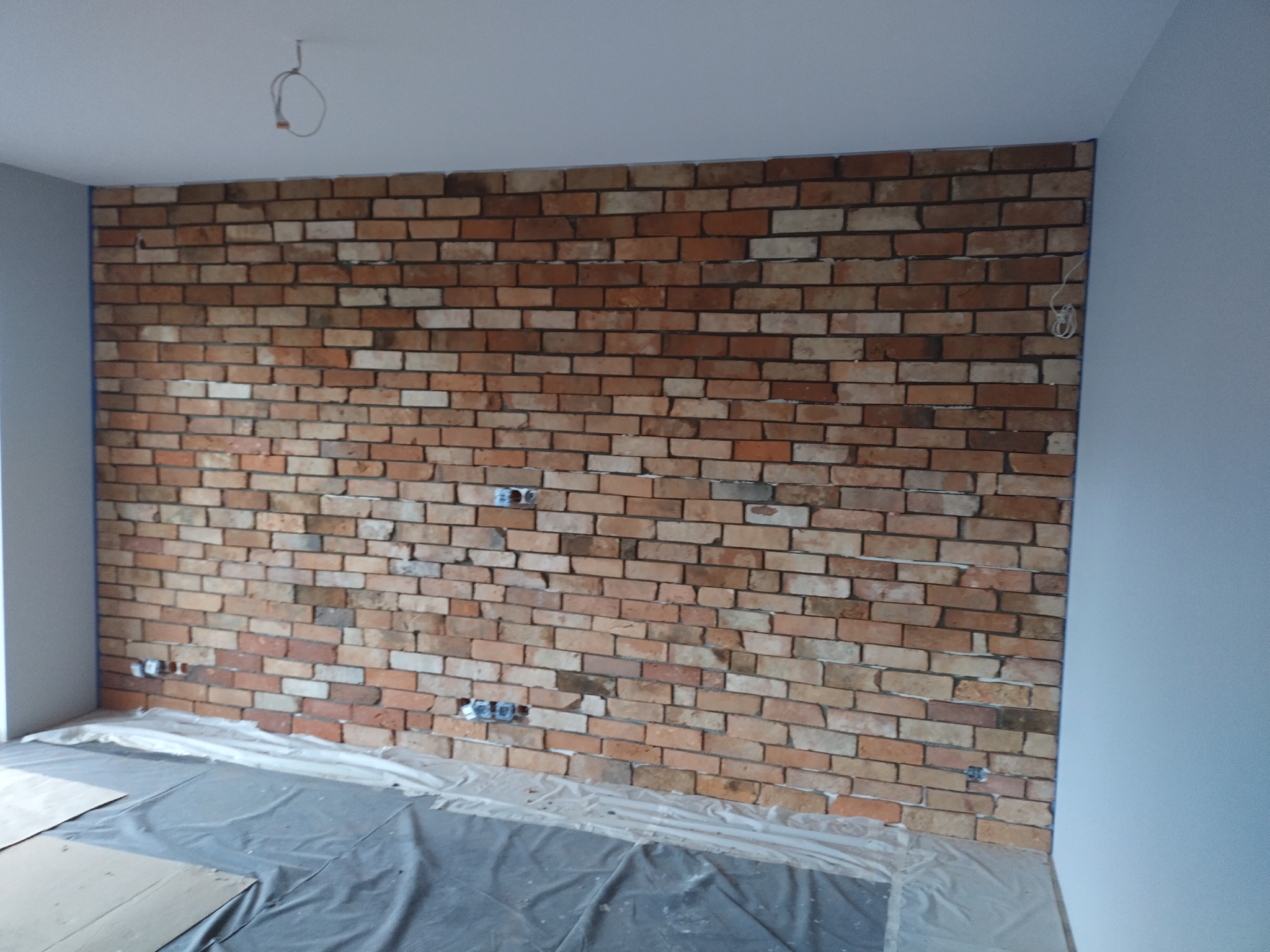 Ściana z cegły klinkierowej w trakcie montażu, widoczne puszki elektryczne, podłoga zabezpieczona folią malarską, niebieska taśma malarska na krawędzi ściany.