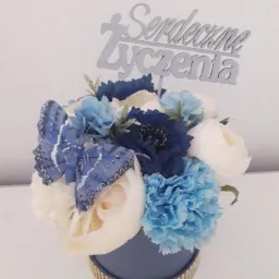 Dekoracja kwiatowa w niebiesko-białej kolorystyce, z papierowymi motylami i napisem 'Serdeczne Życzenia' na srebrnym topperze, umieszczona w niebieskim pudełku z ozdobną, błyszczącą taśmą.