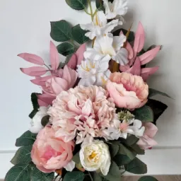 Kompozycja sztucznych kwiatów w odcieniach różu, bieli i zieleni w czarnym wazonie, ustawiona na drewnopodobnej powierzchni pod białą ścianą.