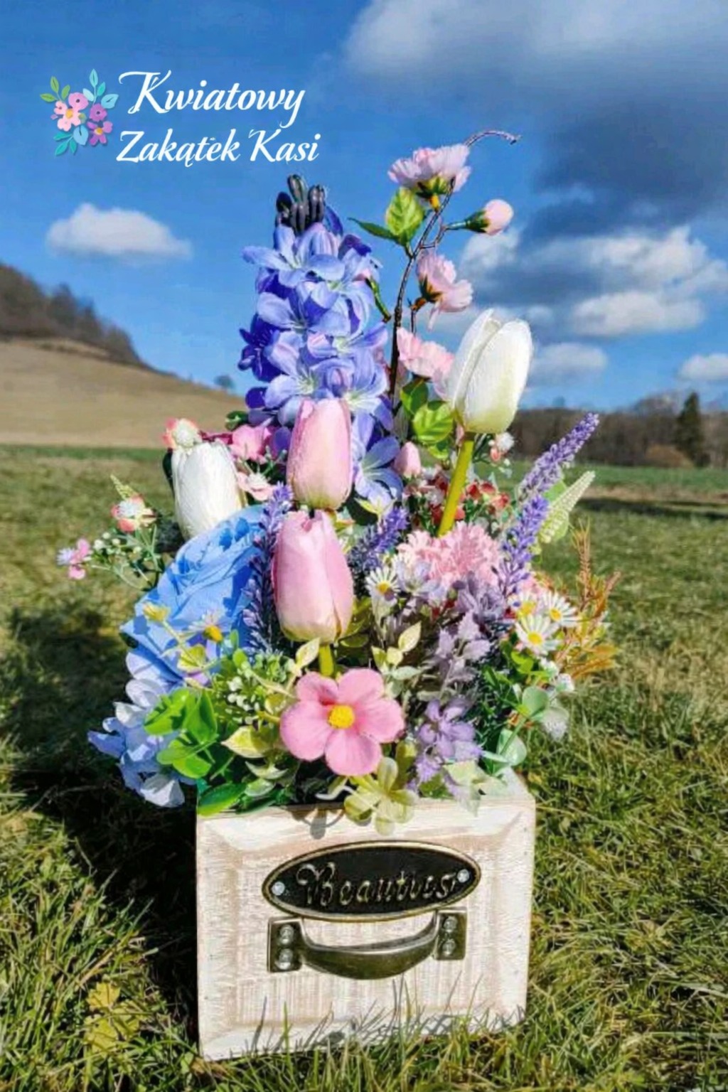 Kompozycja kwiatowa w drewnianej skrzynce z uchwytem, na tle łąki i błękitnego nieba. Różowe tulipany, niebieskie hiacynty i białe tulipany tworzą barwny bukiet.