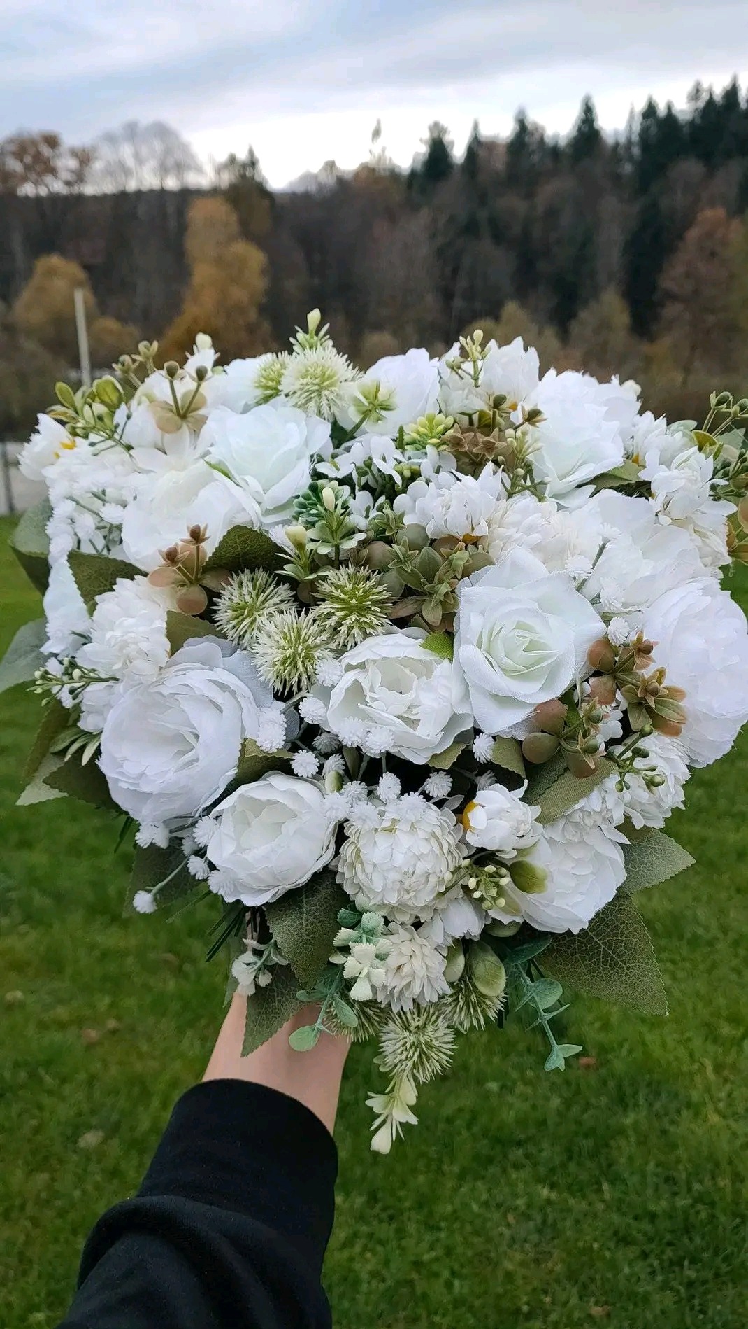 Delikatny, biały bukiet ślubny z róż i zielonych dodatków, trzymany w dłoni na tle zielonego trawnika i drzew. Idealny na eleganckie przyjęcie.