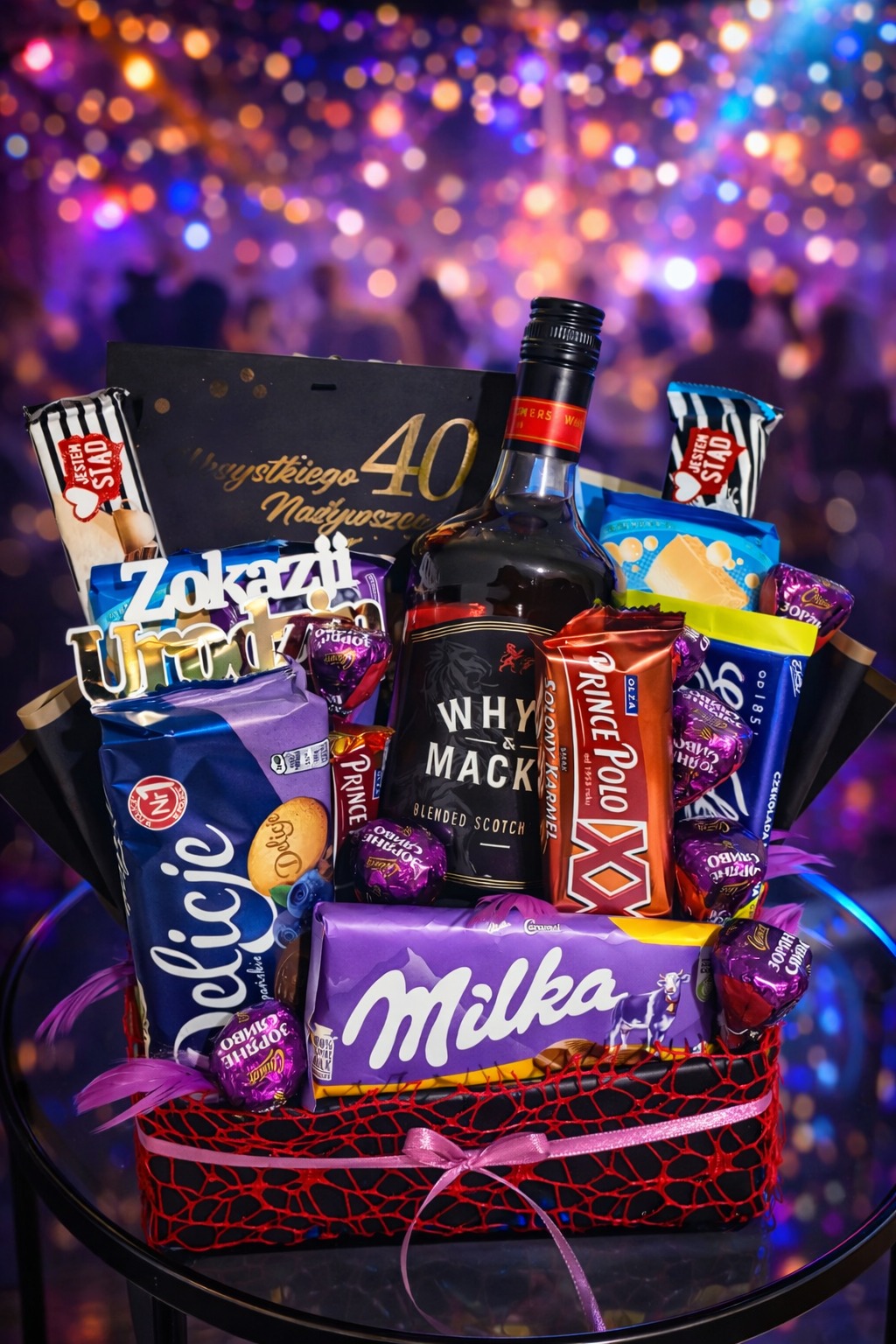Kosz prezentowy na 40. urodziny: whisky, słodycze (Milka, Prince Polo, Delicje) i dekoracje na tle rozświetlonej sceny. Idealny upominek na imprezę.