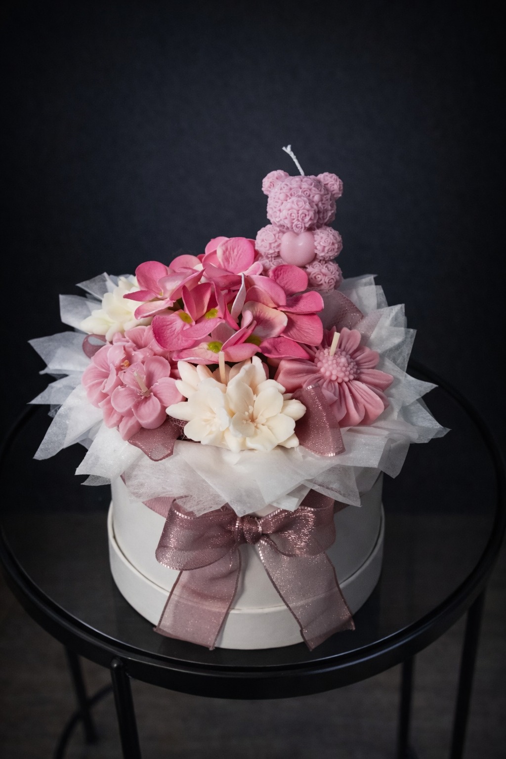 Ręcznie robiony bukiet z mydła w pudełku, z różowymi i białymi kwiatami oraz świeczką w kształcie misia, przewiązany błyszczącą wstążką.