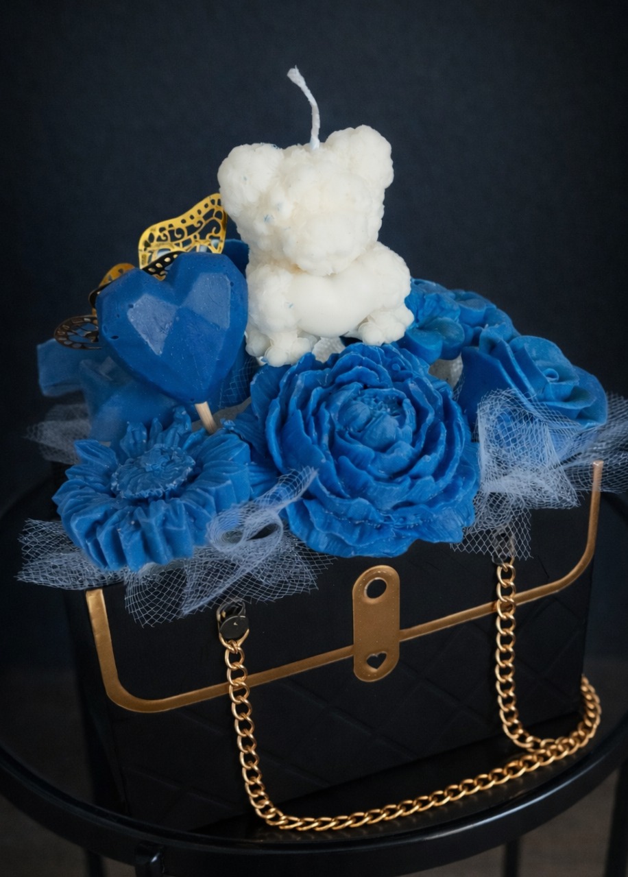 Ręcznie robiona kompozycja świec: biały miś, niebieskie róże i serce w czarnej torebce z złotym łańcuszkiem, na ciemnym tle. Dekoracja stołu na imprezę.