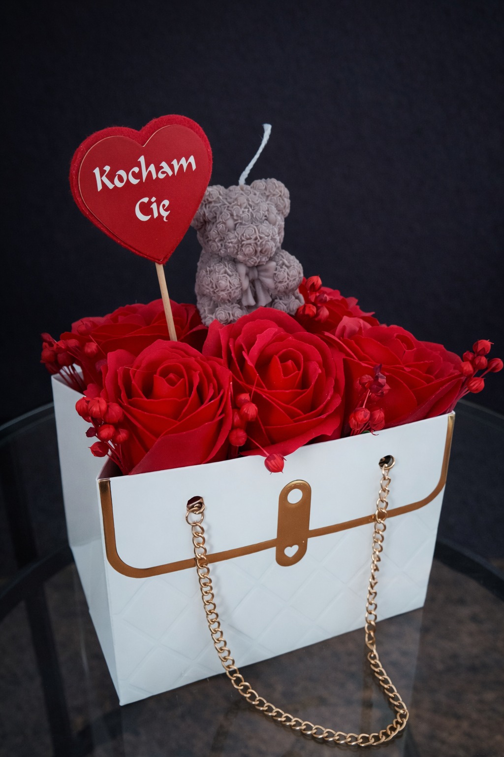Czerwone róże w białej torebce z napisem 'Kocham Cię' i świeczką w kształcie misia. Romantyczny upominek na walentynki lub inną okazję.