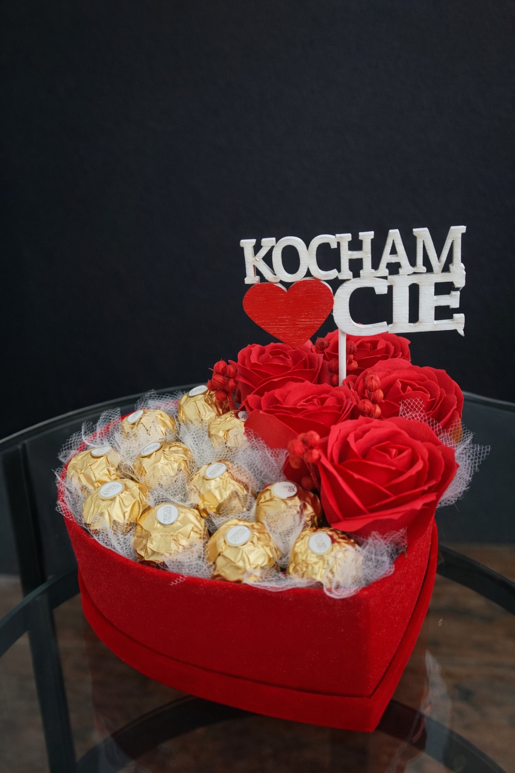 Czerwone pudełko w kształcie serca wypełnione różami, czekoladkami Ferrero Rocher i napisem 'Kocham Cię', na szklanym stole i czarnym tle.