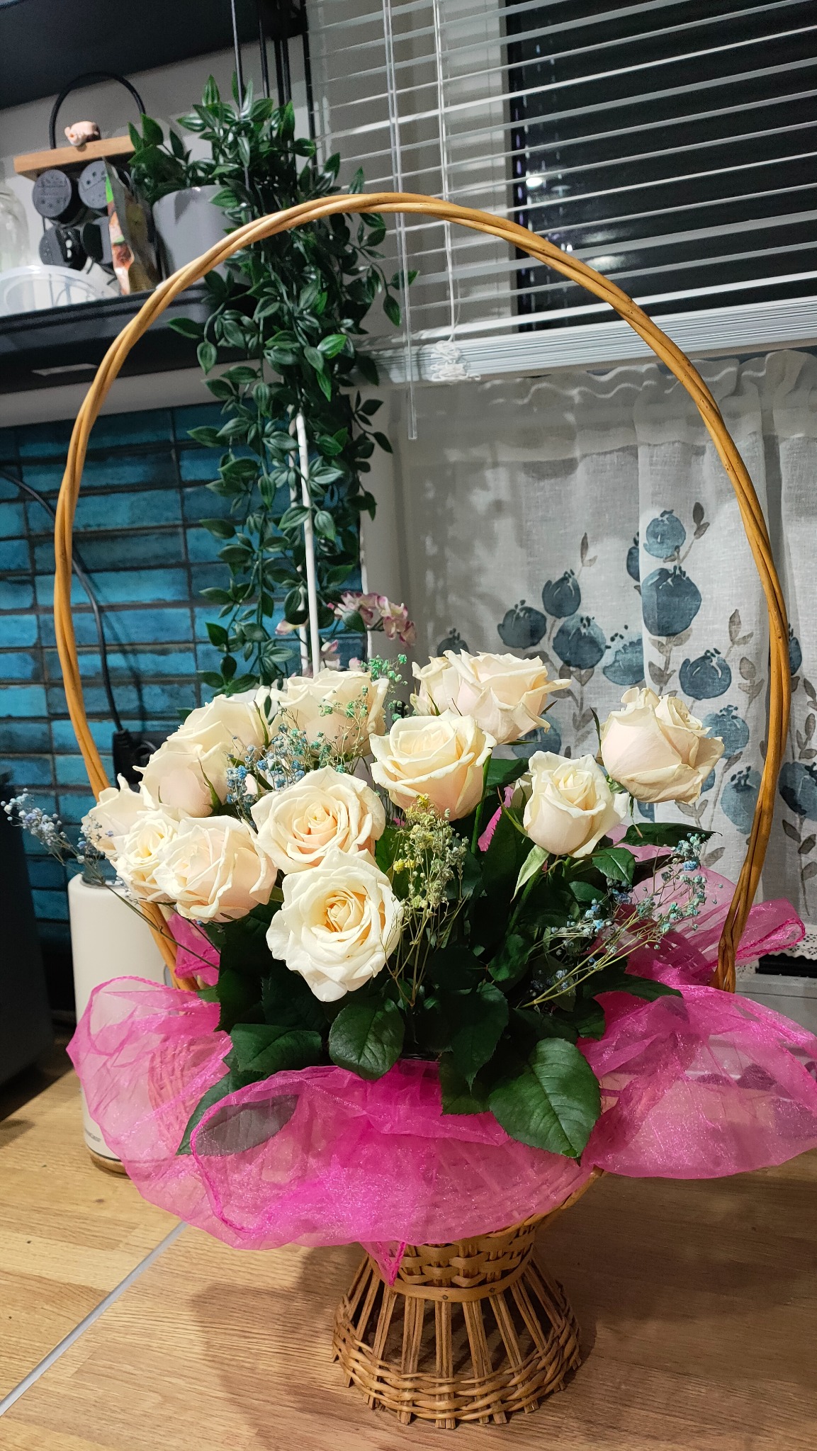 Kompozycja z kremowych róż w okrągłym, wiklinowym koszu, ozdobiona różową tkaniną i drobnymi kwiatami, na tle okna z zasłonami i roślinami.