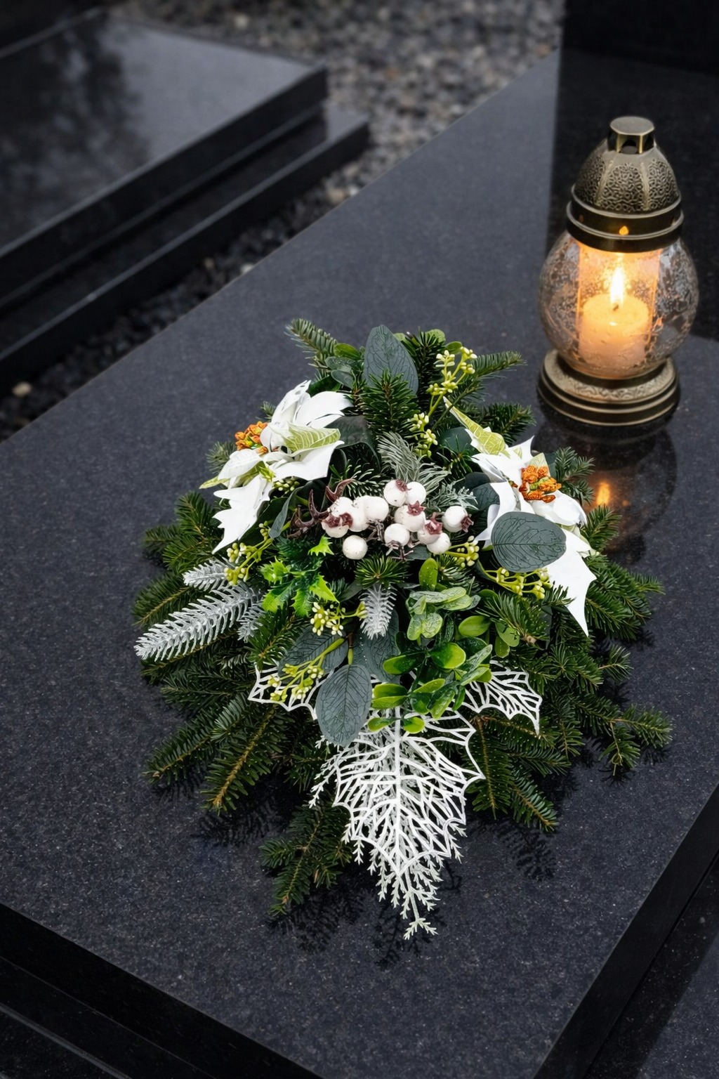 Dekoracja nagrobna z białymi kwiatami i lampionem na czarnym, błyszczącym nagrobku. Kompozycja świąteczna z elementami zieleni i srebrnych akcentów.