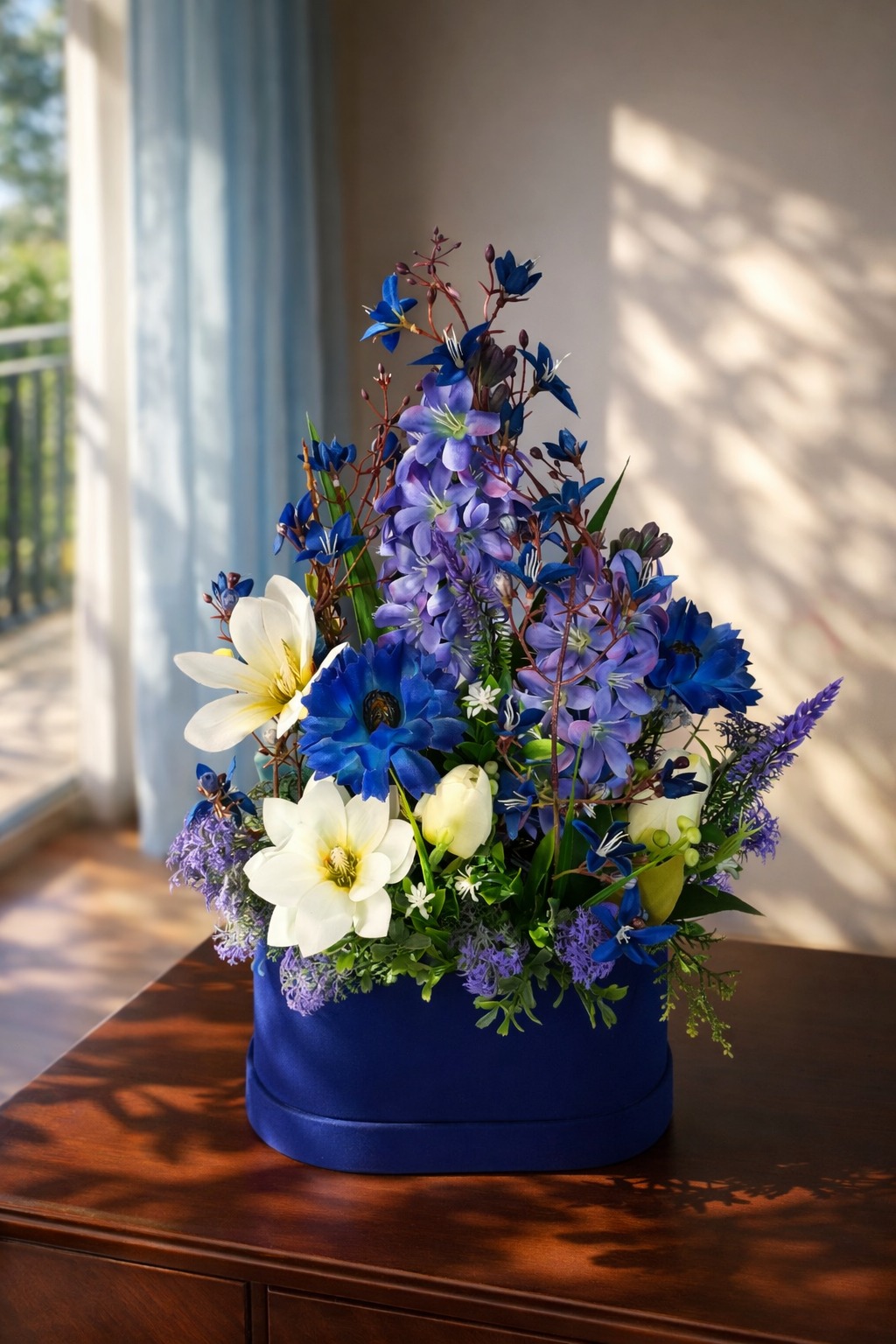 Kompozycja kwiatowa w niebieskim pudełku na drewnianym stole, z widokiem na zewnątrz przez okno z delikatną firanką i padającym cieniem.