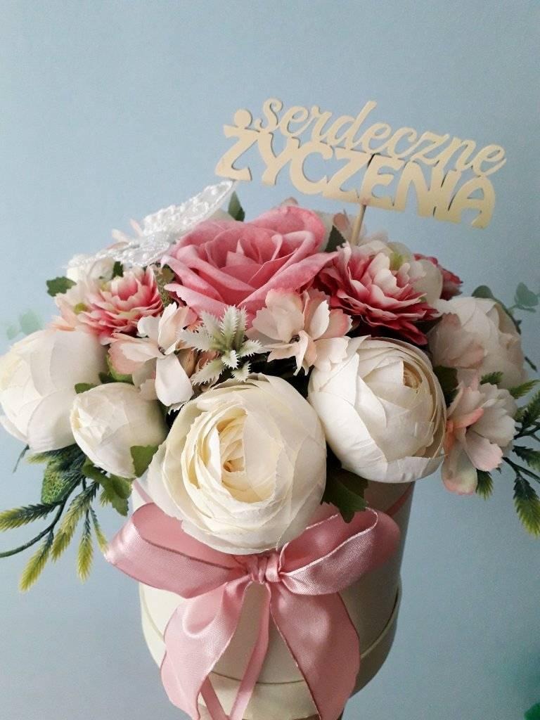 Kompozycja kwiatowa w okrągłym pudełku z różową wstążką, z napisem 'Serdeczne Życzenia' na drewnianym topperze, na tle jasnoniebieskiej ściany.