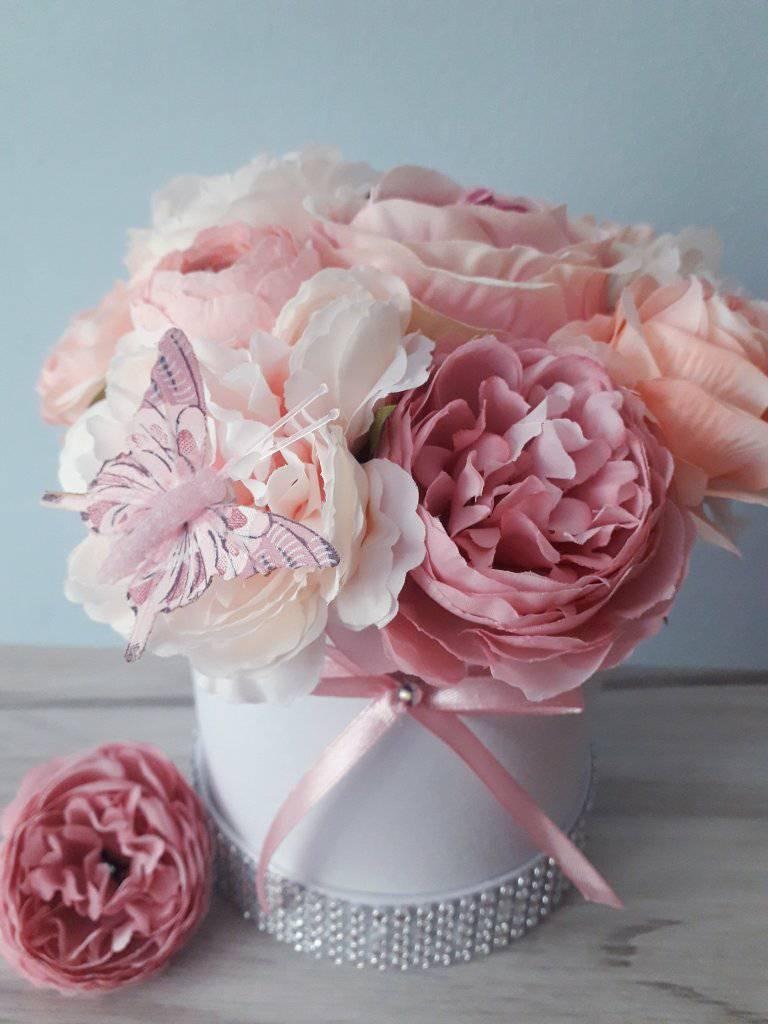 Kompozycja kwiatowa w białym pudełku z różową wstążką i ozdobnym motylem, z dodatkowym kwiatem obok pudełka, na tle błękitnej ściany.