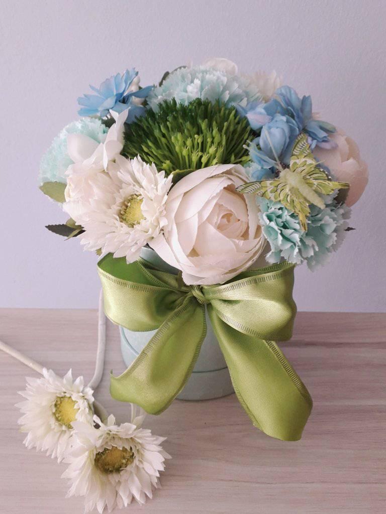 Dekoracyjny bukiet z sztucznych kwiatów w pastelowych odcieniach błękitu, bieli i zieleni, umieszczony w okrągłym, jasnoniebieskim pudełku przewiązanym zieloną satynową wstążką, z dwoma dodatkowymi...