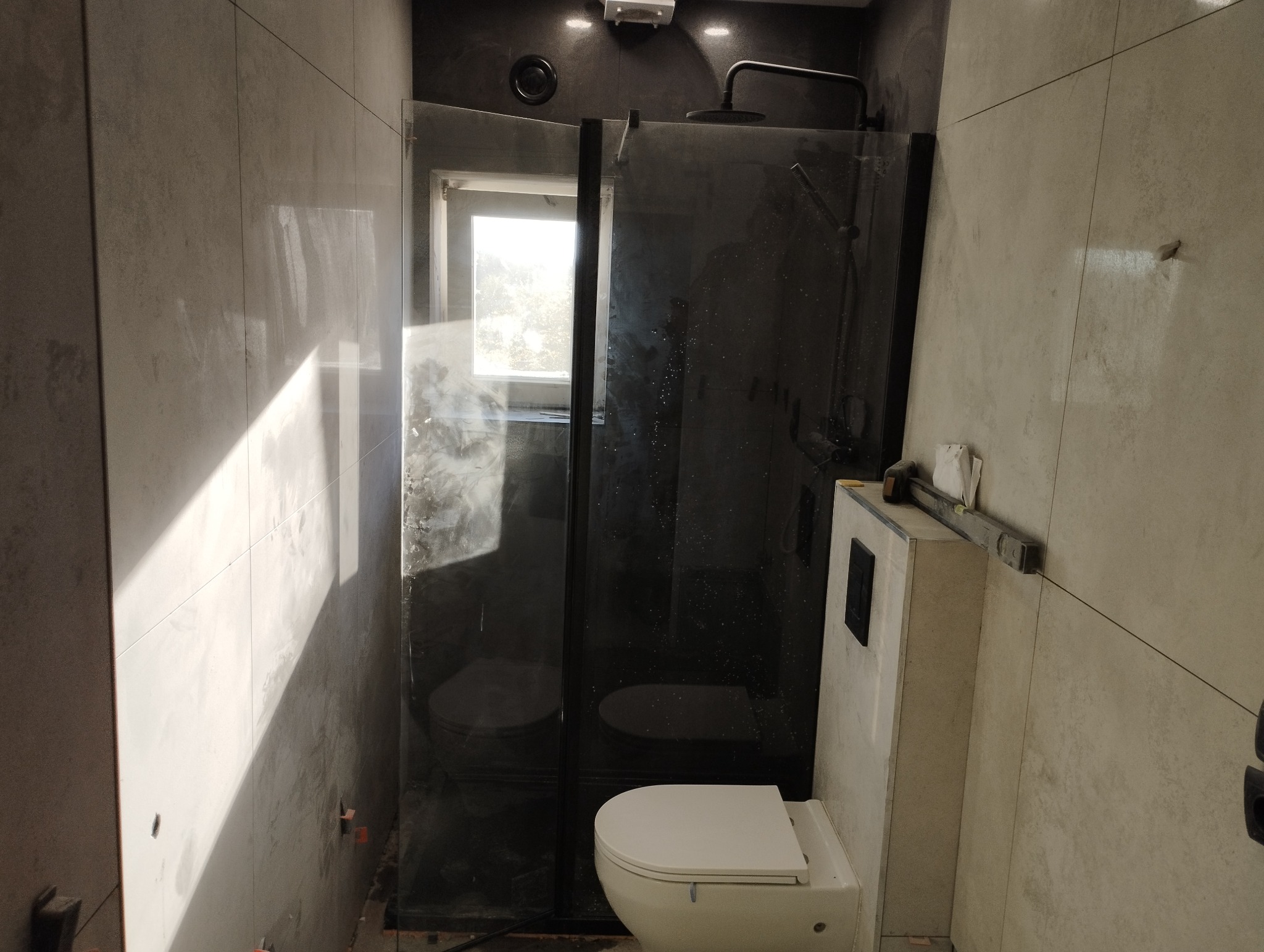 Nowoczesna kabina prysznicowa z czarnymi profilami i deszczownicą, widoczna toaleta w zabudowie, szare płytki ścienne, poziomica na zabudowie toalety.