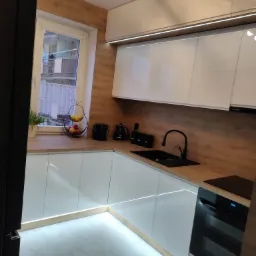 Nowoczesna kuchnia z białymi, lakierowanymi frontami, drewnianym blatem i oświetleniem LED podszafkowym oraz przypodłogowym, czarna armatura i sprzęt AGD.