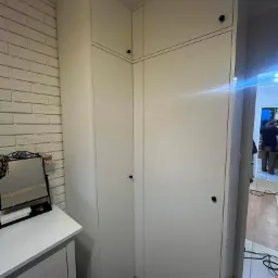 Biała szafa narożna z czarnymi gałkami, dopasowana do ściany z imitacji cegły, obok komoda z lustrem i widoczne kable na podłodze.