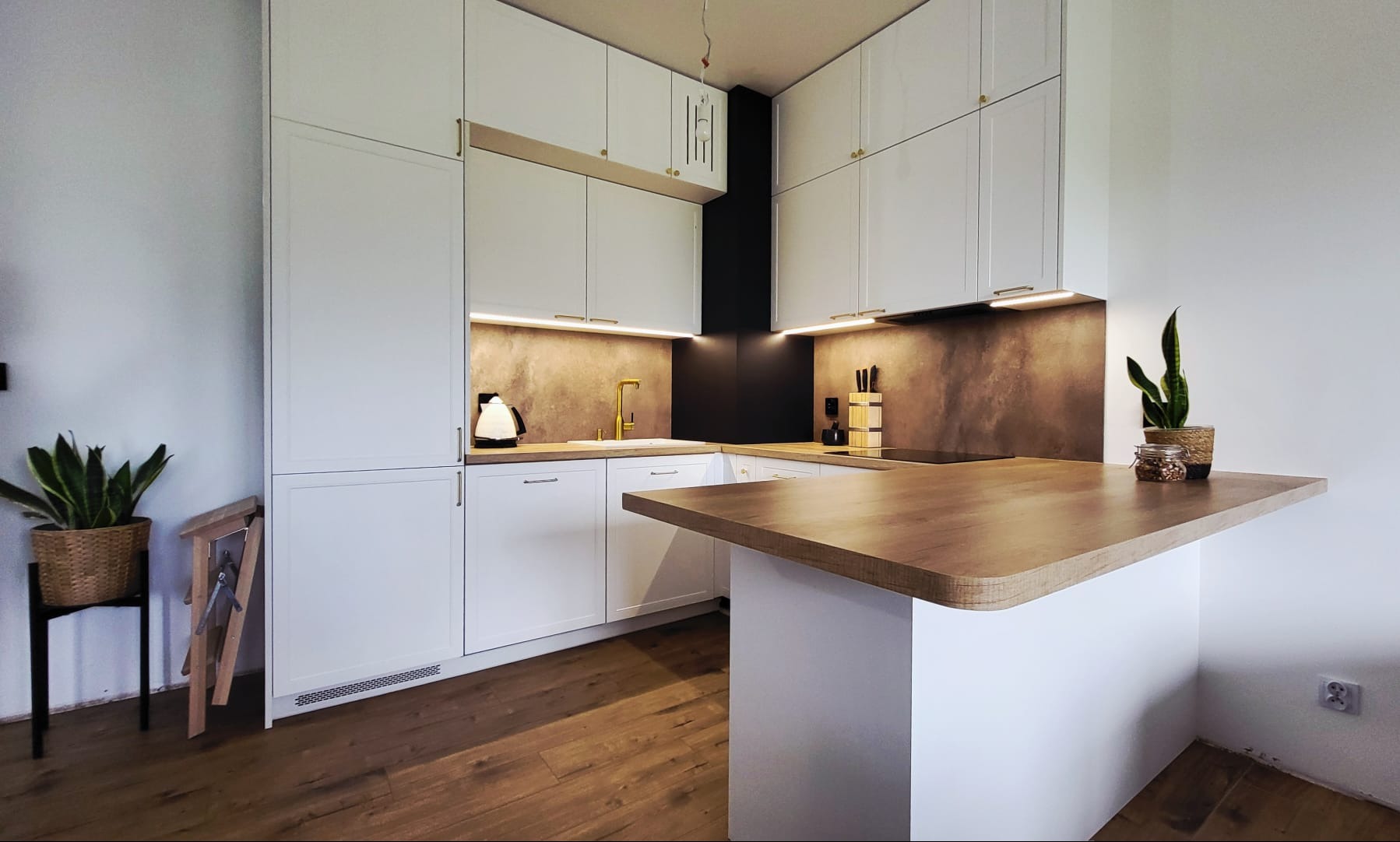 Nowoczesna kuchnia na wymiar z białymi szafkami, drewnianym blatem i złotymi akcentami, oświetlona ciepłym światłem LED, z roślinami doniczkowymi jako element dekoracyjny.