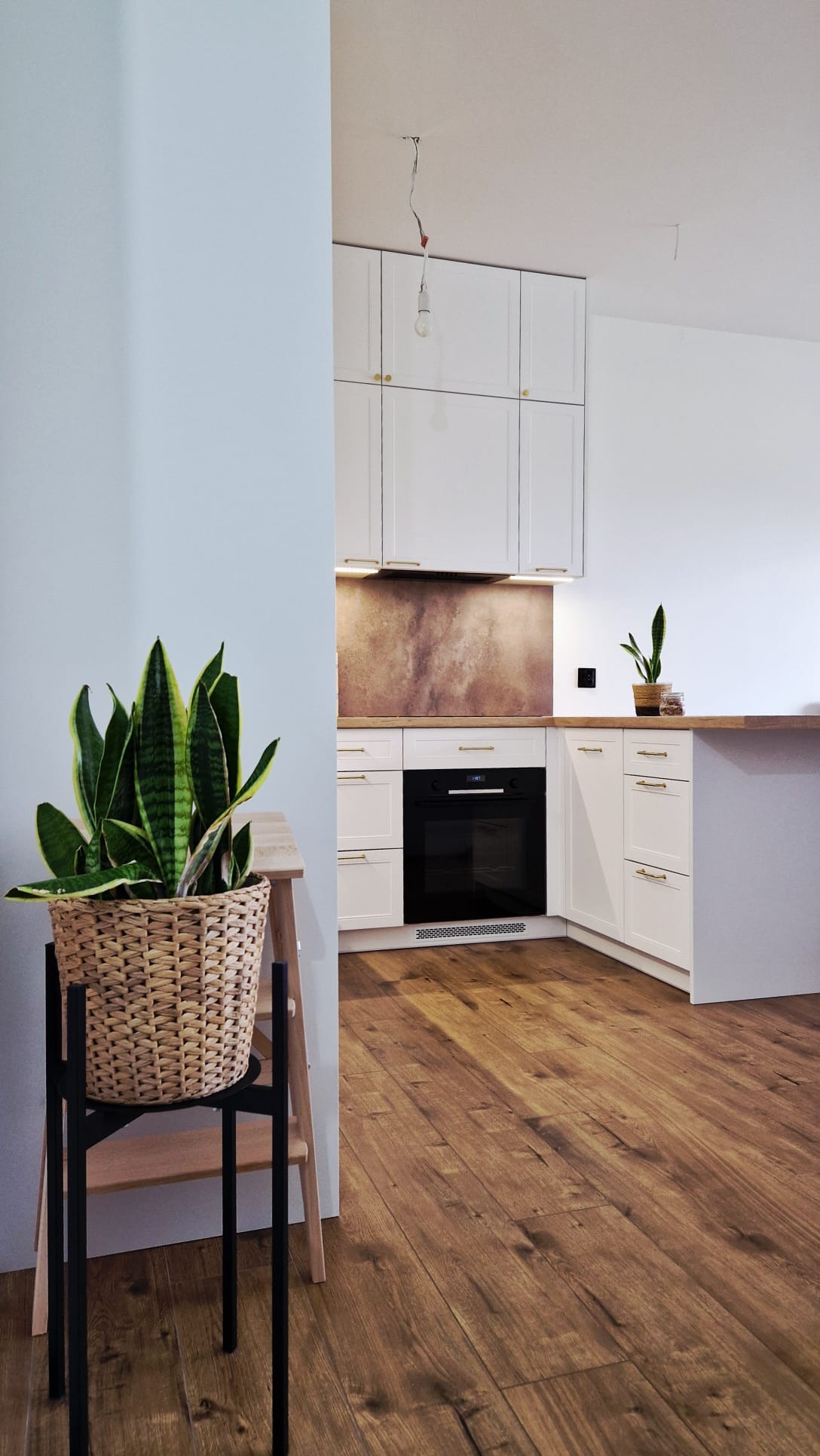 Jasna kuchnia z białymi szafkami i drewnianym blatem, widziana z perspektywy salonu. Na pierwszym planie roślina w plecionym koszu na metalowym stojaku.