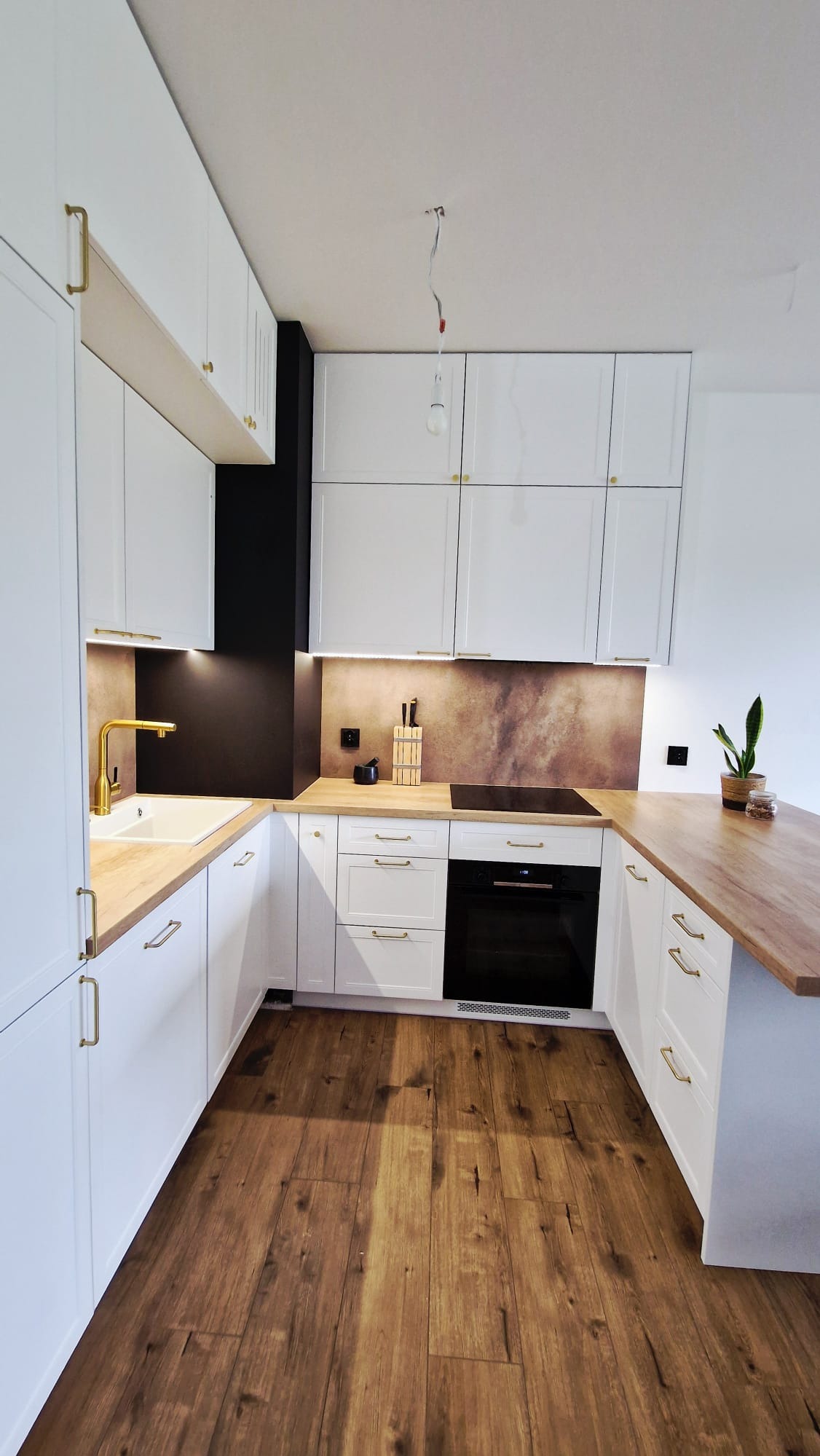 Nowoczesna kuchnia z białymi frontami, złotymi uchwytami i drewnianym blatem. Uwagę przyciąga czarny element nad płytą grzewczą i drewniana podłoga.