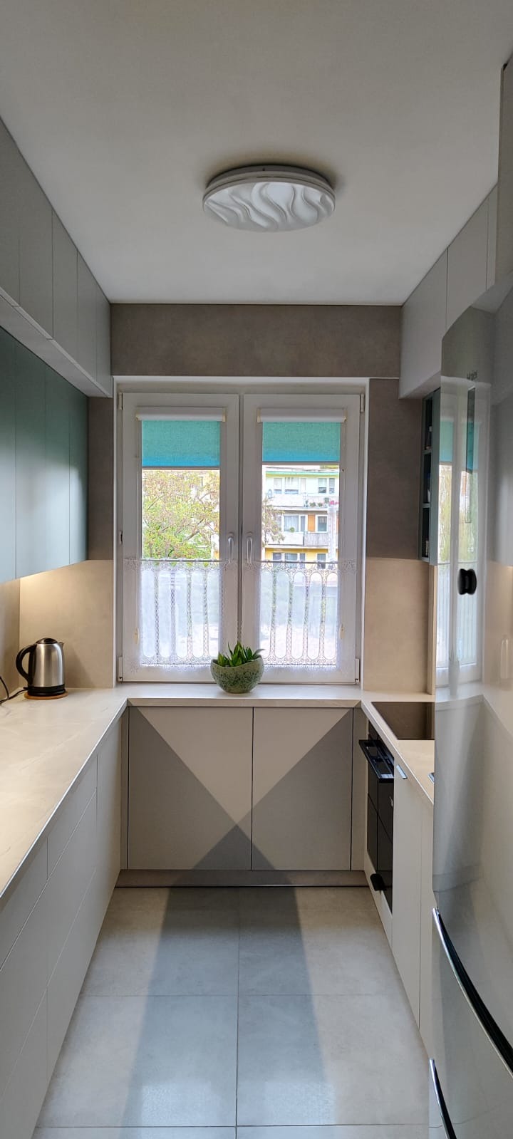 Wnętrze kuchni z oknem, jasnymi szafkami z geometrycznym wzorem, blatem i zielonym akcentem roślinnym. Widok na zewnątrz przez okno.