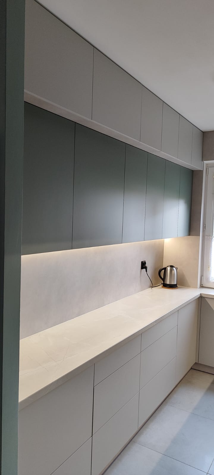Nowoczesna kuchnia z szarymi szafkami górnymi i dolnymi, białym blatem i subtelnym oświetleniem LED pod szafkami. Na blacie czajnik elektryczny.