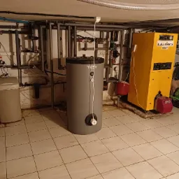 Szary, pionowy bojler TESY z widocznym termostatem, obok żółty piec LUMO, instalacja rur na ścianie i podłodze z jasnych płytek w piwnicy.