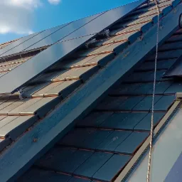 Ciemna dachówka ceramiczna z zamontowanymi oknami dachowymi i panelami fotowoltaicznymi w słoneczny dzień.