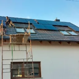 Dwóch monterów instaluje panele słoneczne na dachu domu jednorodzinnego z wykorzystaniem rusztowania; widoczne okna dachowe i szara dachówka.