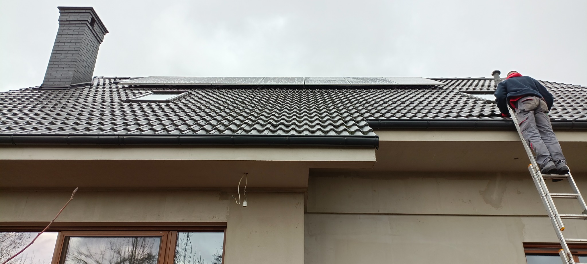 Pracownik na drabinie przy dachu z dachówką ceramiczną, widoczne okna dachowe, komin z cegły klinkierowej oraz panele słoneczne na dachu.