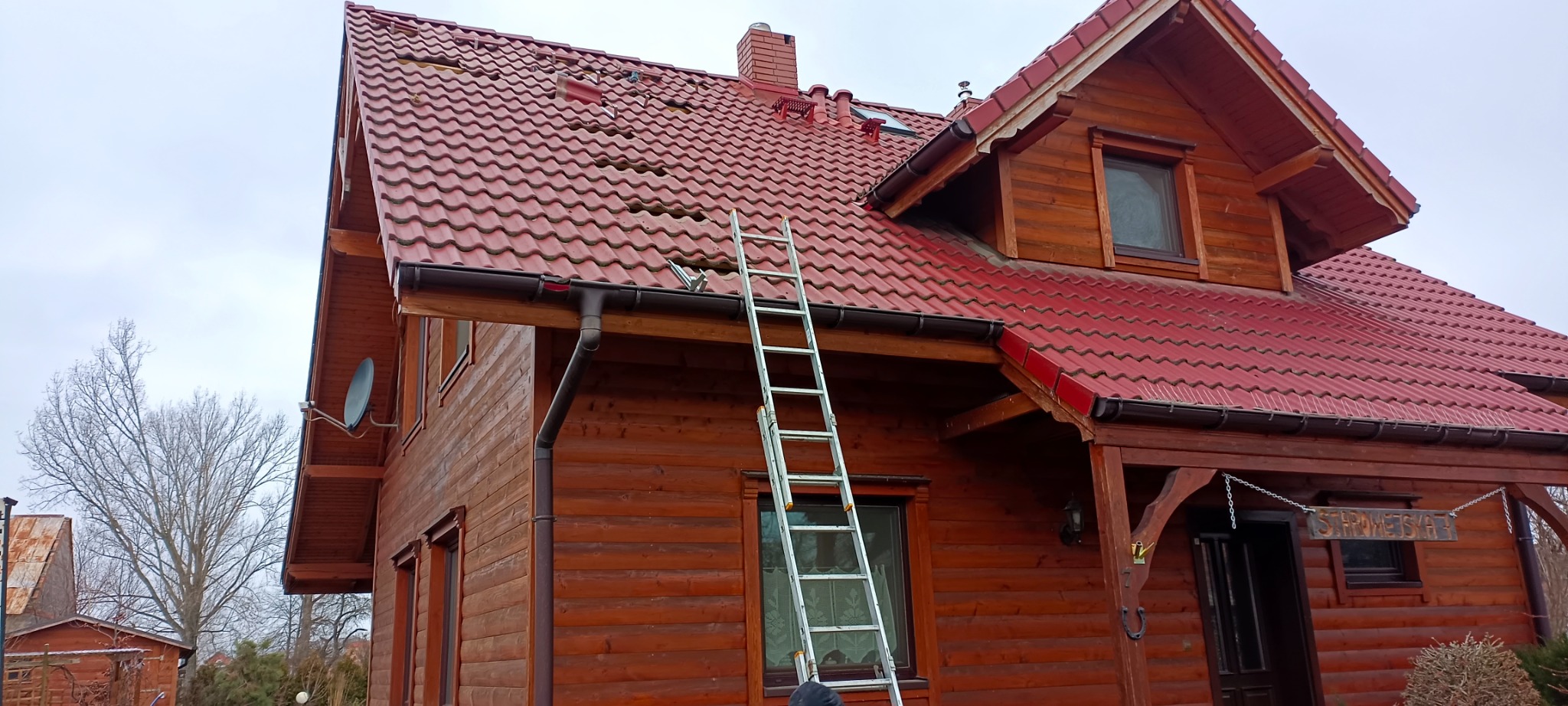 Częściowy widok drewnianego domu z czerwoną dachówką, na której widoczne są ubytki, z przystawioną aluminiową drabiną i satelitą na ścianie.