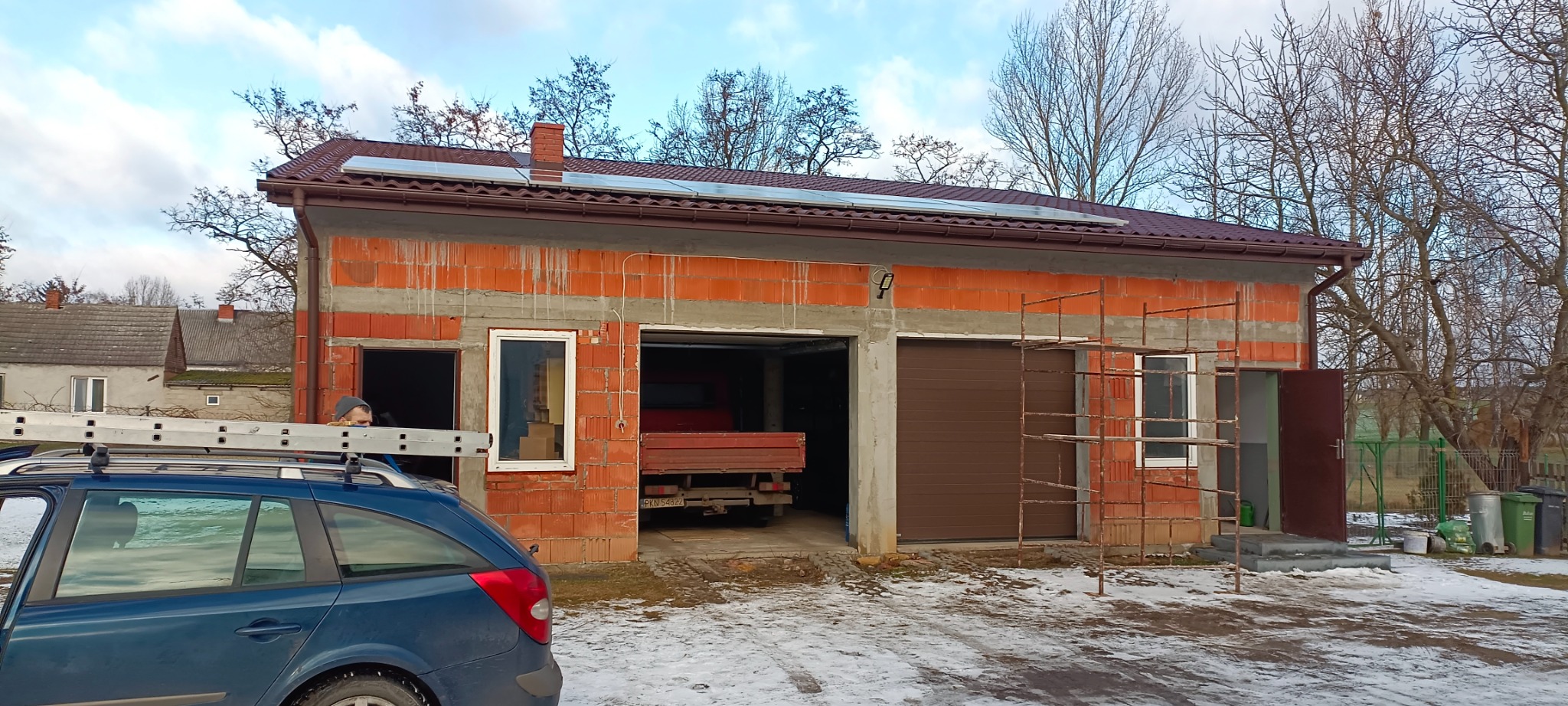 Budynek garażowy w trakcie budowy z czerwonej cegły, z brązowym dachem i panelami fotowoltaicznymi, częściowo otynkowany, ze stalowymi rusztowaniami, na dachu samochodu drabina, w tle zabudowa...
