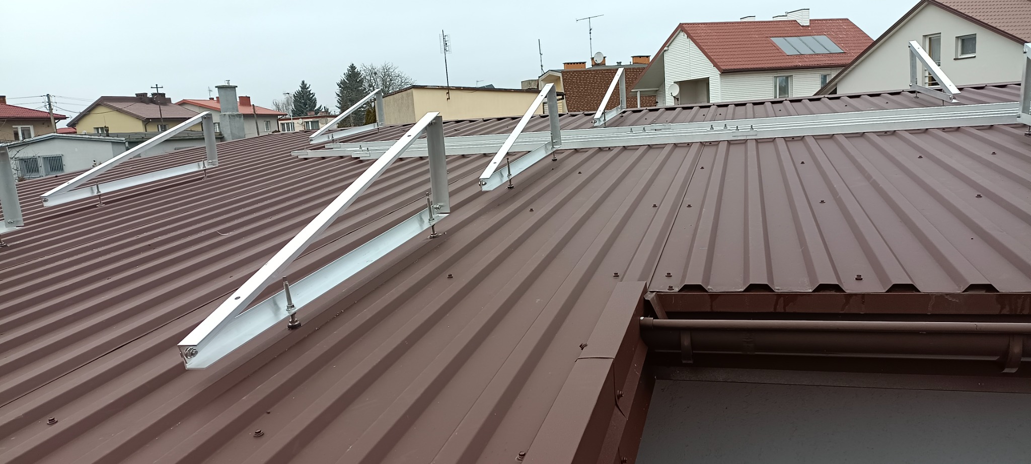 Montaż aluminiowej konstrukcji wsporczej pod panele fotowoltaiczne na brązowym, blaszanym dachu domu jednorodzinnego w pochmurny dzień.