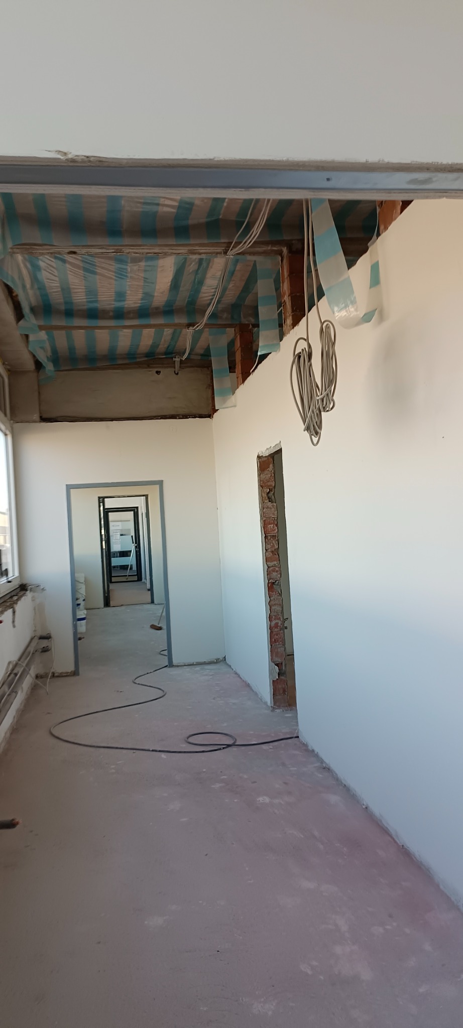 Wnętrze w trakcie remontu: białe ściany, odkryta cegła wokół otworu drzwiowego, podwieszony sufit zabezpieczony folią malarską w paski, zwisające kable elektryczne i przewód na podłodze.