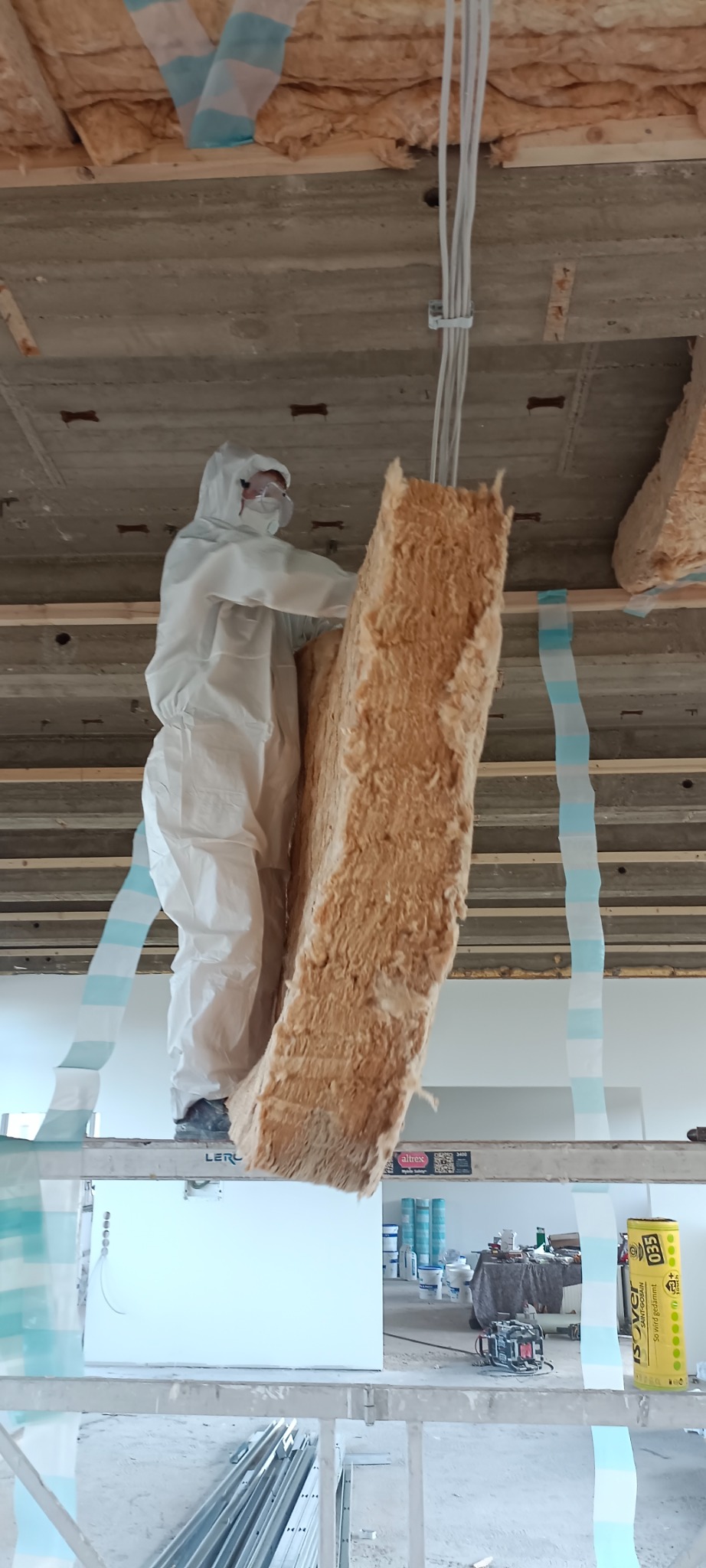 Pracownik w kombinezonie ochronnym montuje izolację z wełny mineralnej na suficie podwieszanym, widoczne drewniane legary i przewody elektryczne