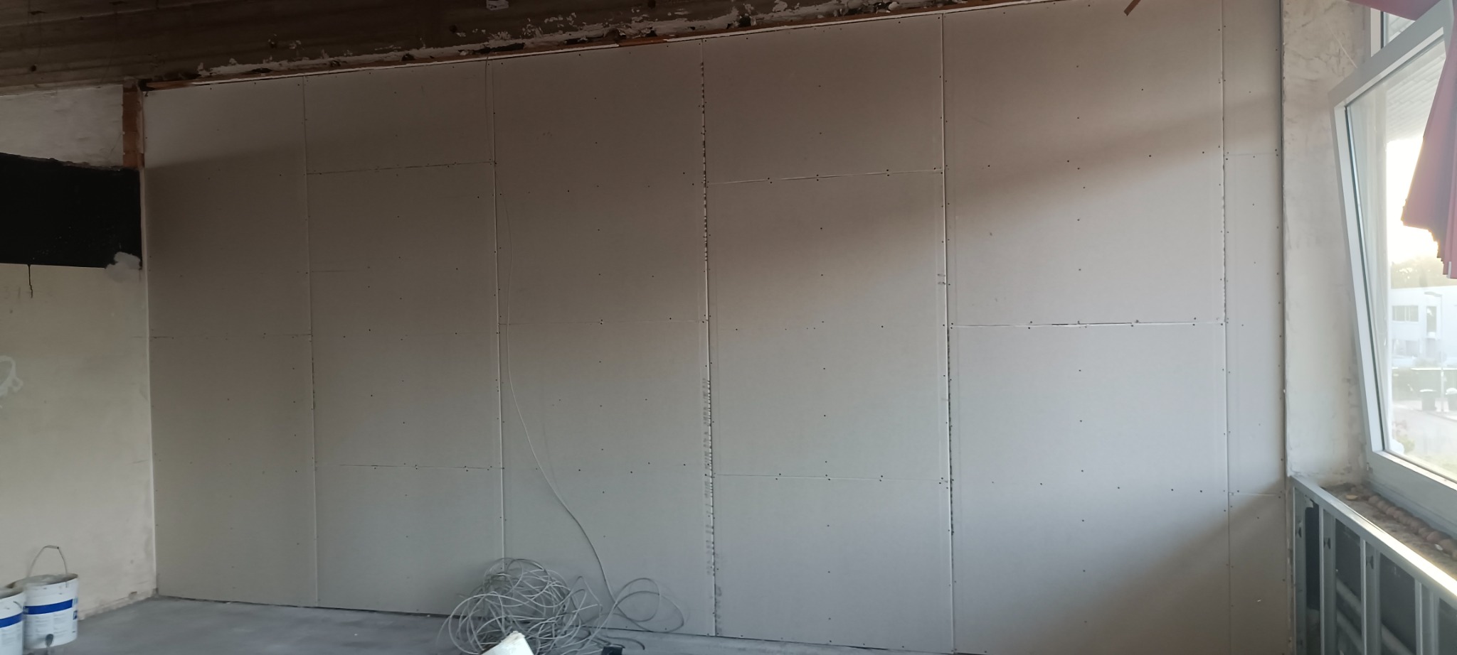 Ściana wewnętrzna wyłożona płytami kartonowo-gipsowymi, widoczne ślady po wkrętach, szpachla i przewody elektryczne na podłodze, fragment okna z widokiem na zewnątrz.