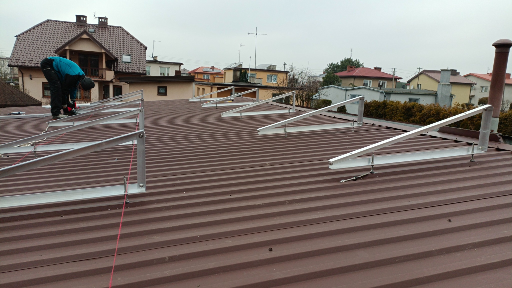 Montaż konstrukcji wsporczej pod panele fotowoltaiczne na brązowym, trapezowym dachu; widoczny pracownik w niebieskiej kurtce przykręcający element konstrukcji, w tle zabudowa jednorodzinna.