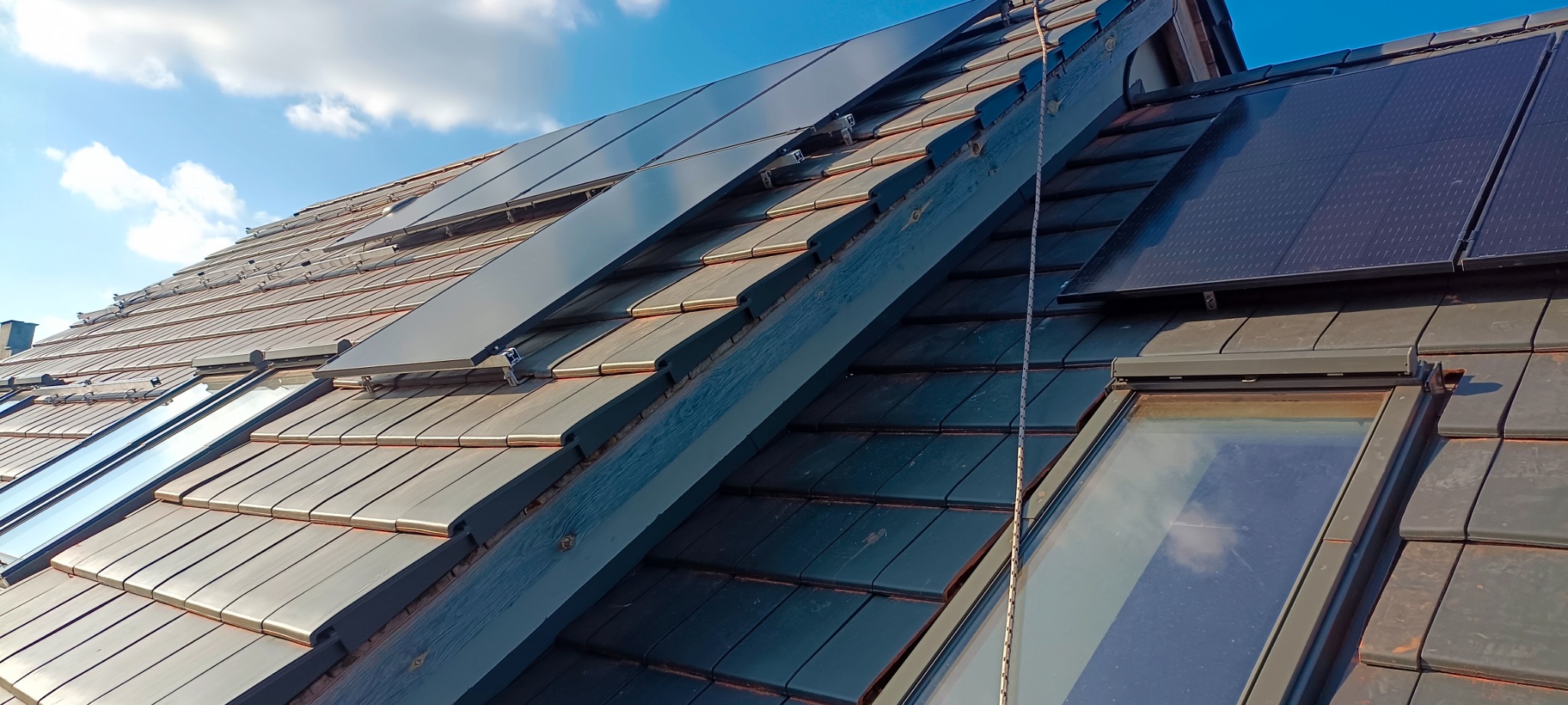 Ciemna dachówka ceramiczna z zamontowanymi oknami dachowymi i panelami fotowoltaicznymi w słoneczny dzień.