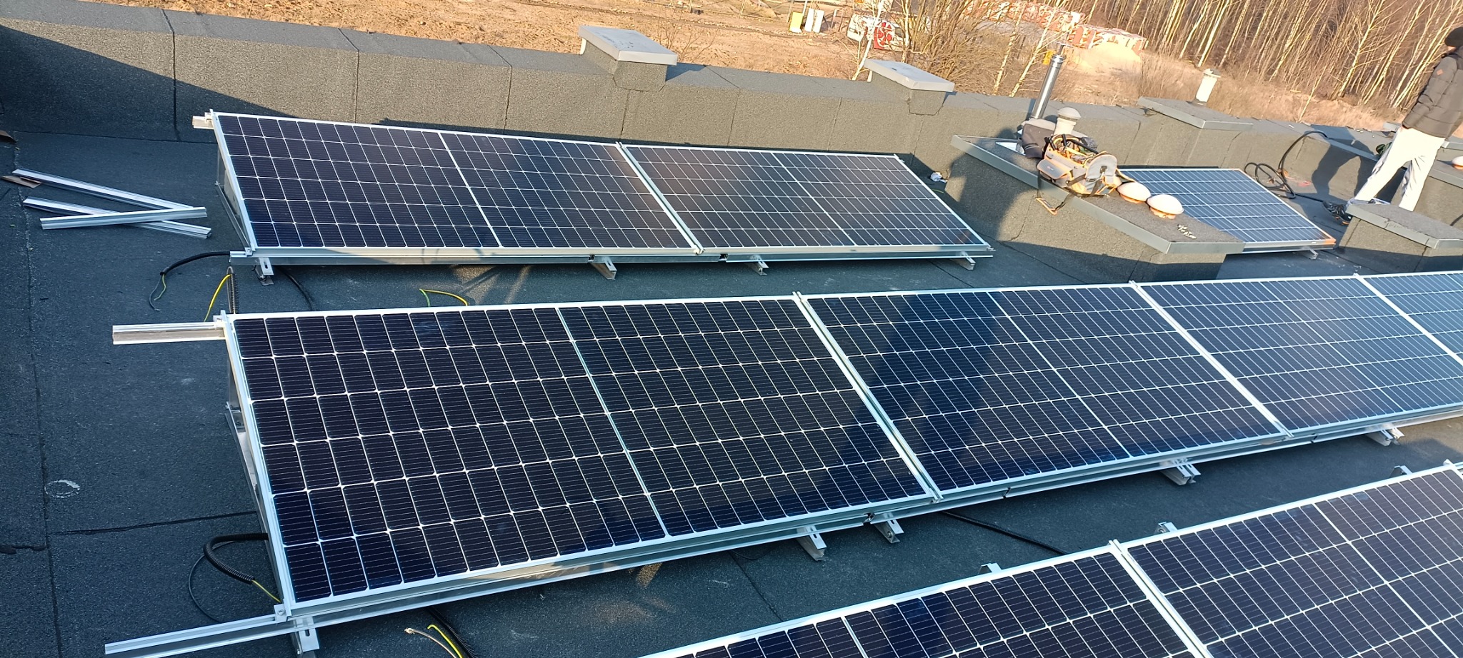 Montaż paneli fotowoltaicznych na płaskim dachu pokrytym papą, widoczne aluminiowe profile montażowe i częściowo ułożone panele oraz narzędzia pracy.