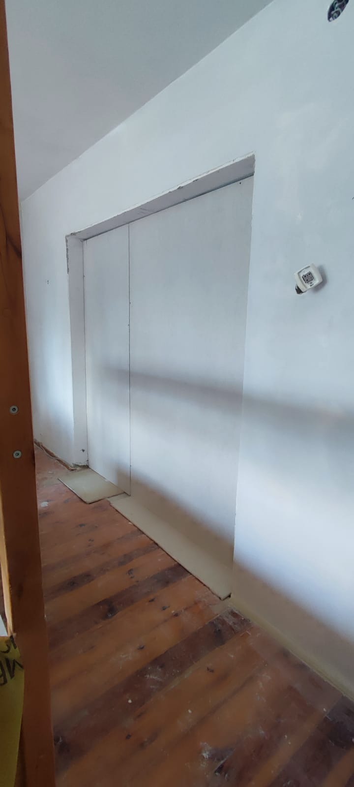 Wnętrze w trakcie prac wykończeniowych: ściany pokryte świeżym szpachlem, widoczna rama bez drzwi, drewniana podłoga częściowo zakryta folią ochronną, gniazdko elektryczne na ścianie.