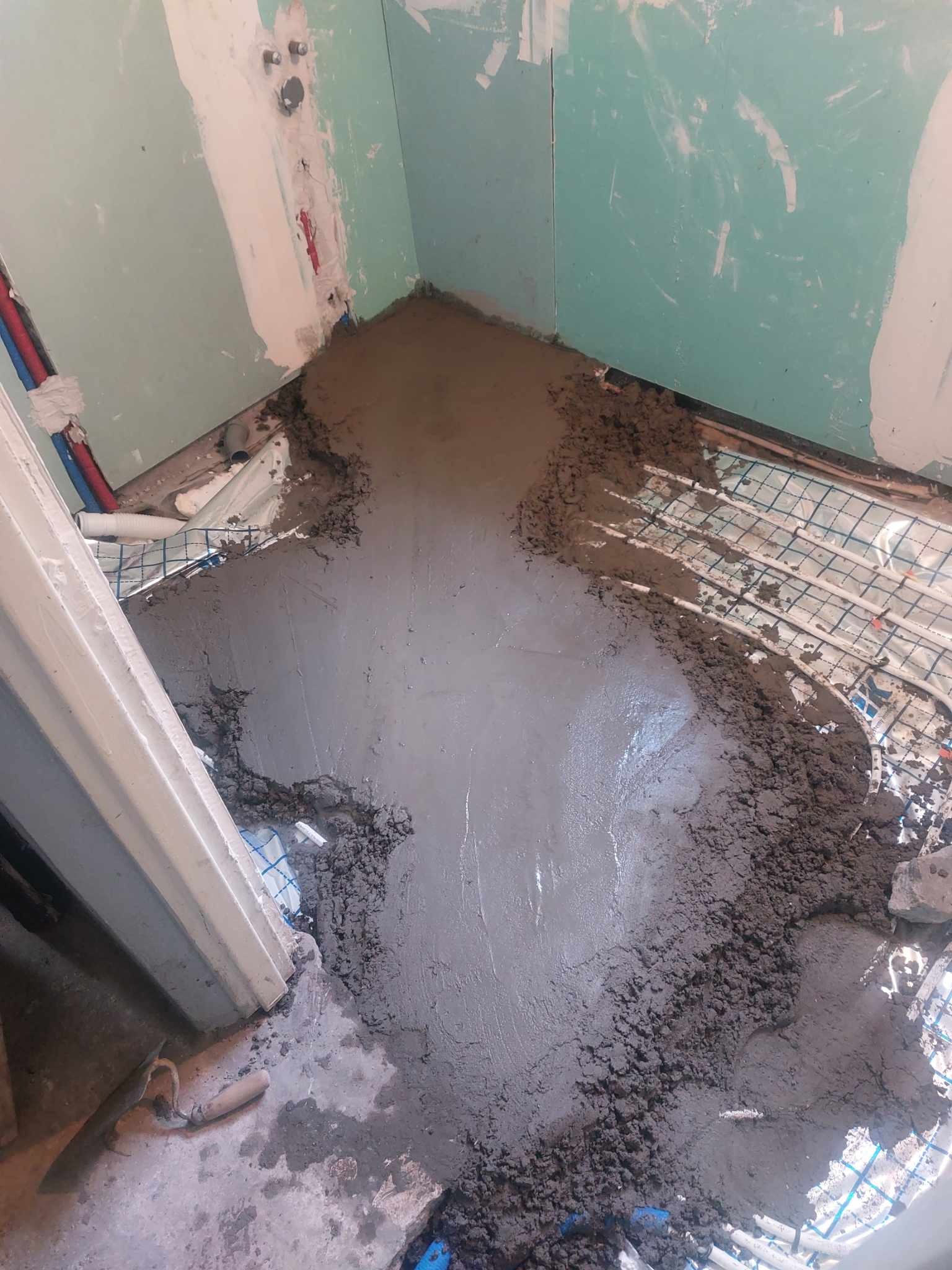 Wylewka betonowa na siatce z ogrzewaniem podłogowym w trakcie prac, widoczne rury instalacji wodnej i fragment ściany z gładzią gipsową.