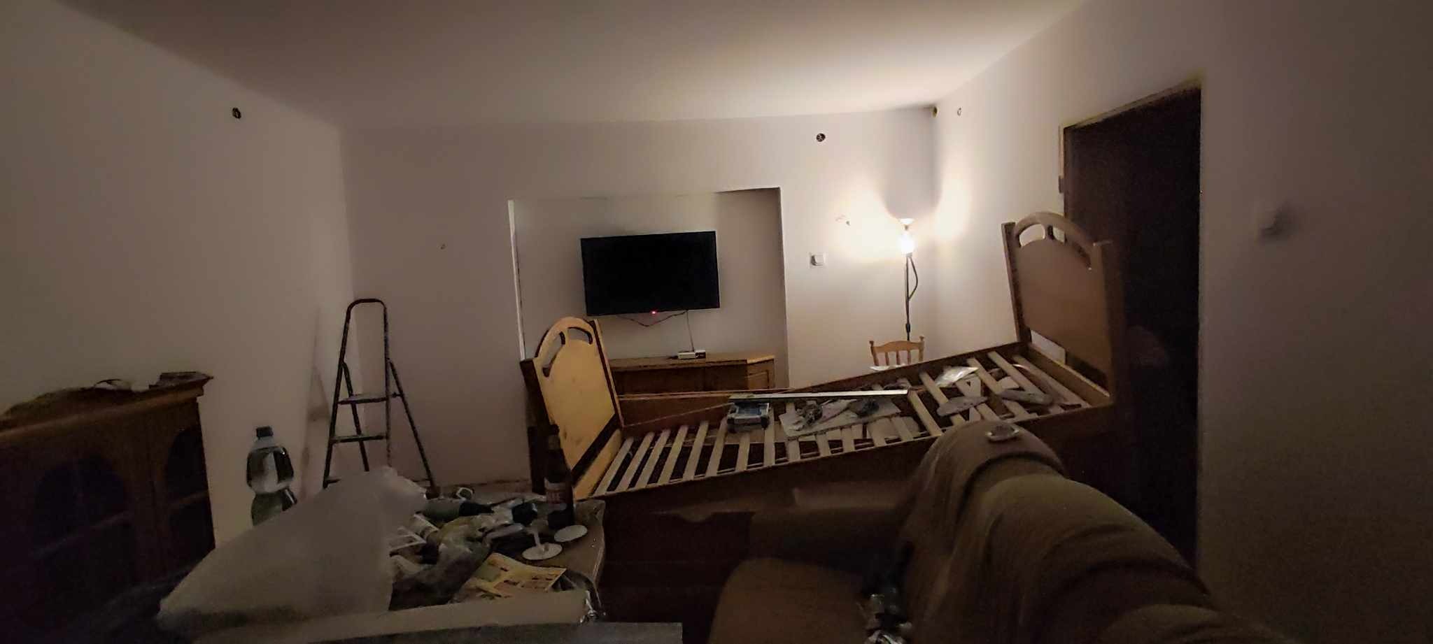 Pokój w trakcie demontażu: rozłożone łóżko, drabina, otwory po kołkach w ścianie, telewizor na komodzie, lampa stojąca.