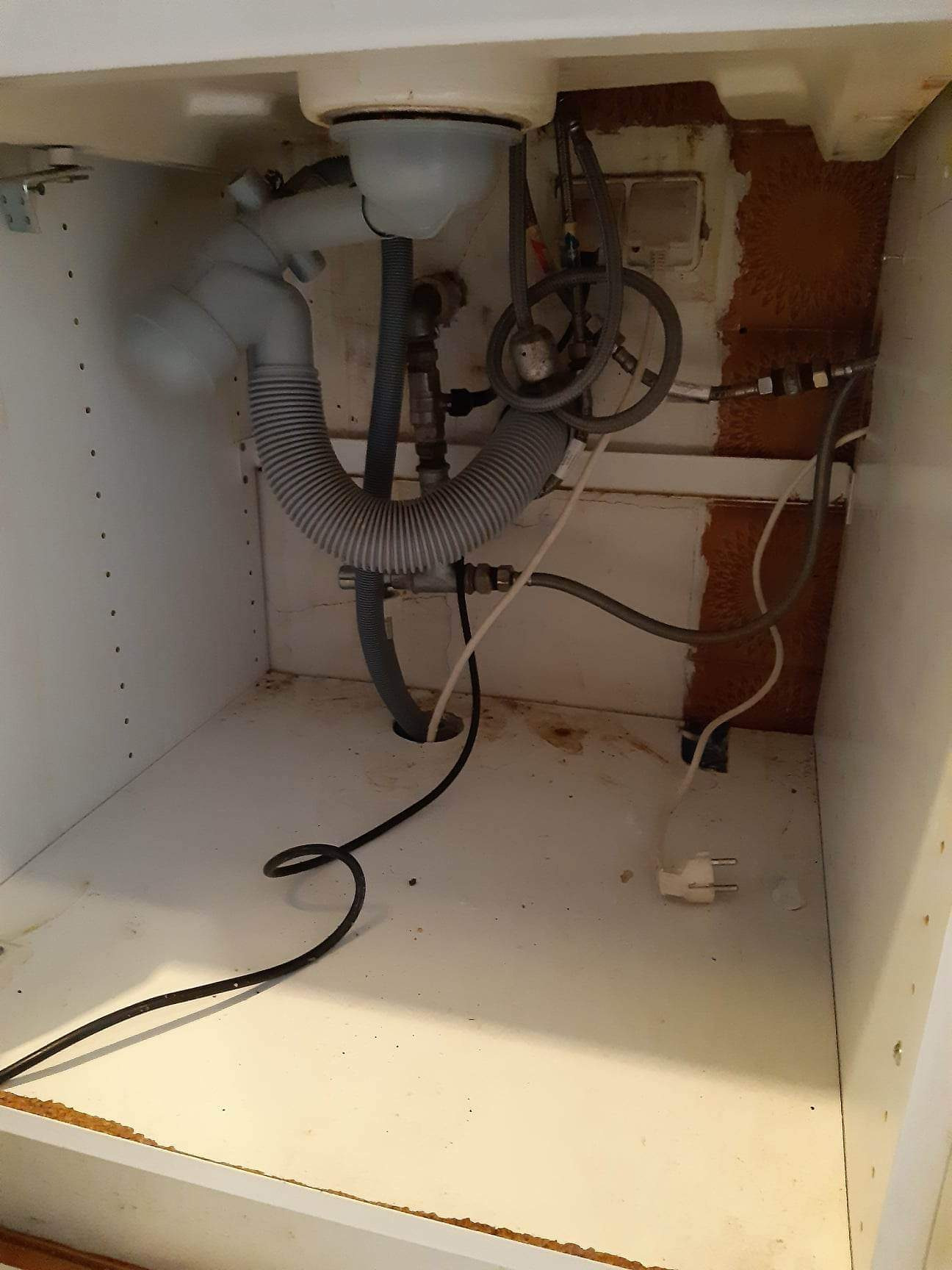 Syfon, rury i zawory pod zlewem w szafce, widoczne ślady użytkowania i zabrudzenia, kabel zasilający z wtyczką w dolnej części kadru.