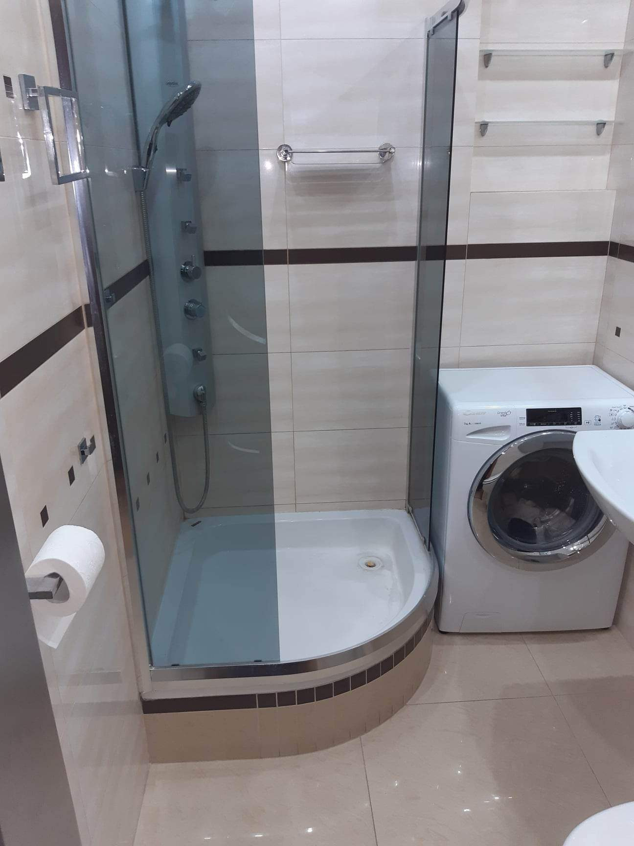 Narożna kabina prysznicowa z hydromasażem, umywalka i pralka w łazience wyłożonej beżowymi płytkami z brązowymi akcentami.