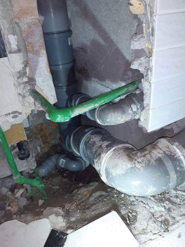 Fragment ściany z odsłoniętymi rurami kanalizacyjnymi z szarego PCV i zielonymi rurami doprowadzającymi wodę, widoczne ślady po kuciu w ścianie i podłodze.