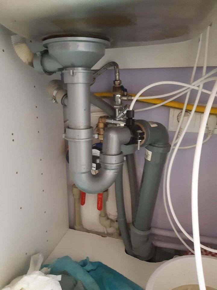 Szare rury odpływowe pod zlewem kuchennym, widoczne zawory odcinające wodę i białe wężyki doprowadzające wodę do filtra.