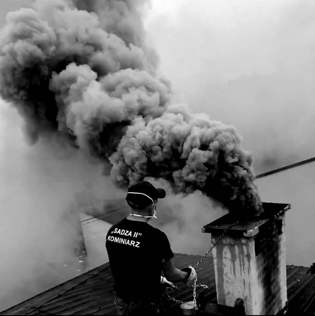Kominiarz w masce i czapce na dachu czyści komin, z którego wydobywa się gęsty, ciemny dym; widoczny napis 'SADZA II' na koszulce.